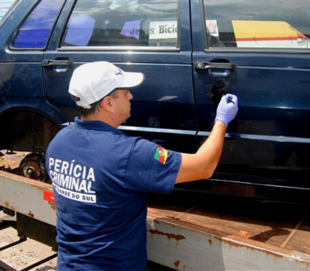 Homem com roupa azul escuro da perícia criminal do Rio Grande do Sul e bone branco realizando a perícia no carro azul marinho 