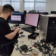 Homem de camiseta preta sentado em frente a um computador com equipamentos a sua volta