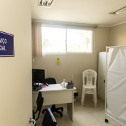 Sala do CRAIs com uma placa azul com os dizeres "Serviço Social" em branco sobre uma porta bege. Dentro da sala é possível ver uma escrivaninha com computador, uma pilha de cadeiras plásticas brancas e duas cadeiras pretas estofadas.