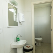 Banheiro branco com uma porta divisória. Antes da porta, há uma pia, um espelho, um porta papel-toalha e um lixo, e depois da porta é visível um vaso sanitário.