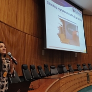 Mulher segurando microfone em um auditório e um painel com informações sobre o tema que ela está palestrando 