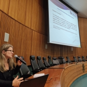 Mulher segurando um microfone em um auditório e um painel ao fundo com algumas informações sobre o tema que ela está palestrando 