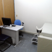 Sala do PML. Mesa com computador em frente a uma cadeira azul e atrás de uma cadeira preta, em um canto entre a parede branca e a divisória de madeira clara. Do outro lado da pequena sala, há uma mesa estofada branca para exames.